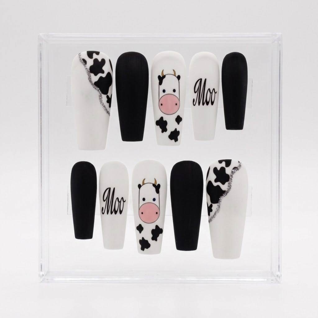 A set of 10 coffin-shaped press-on nails displayed in a clear acrylic case. The nails feature a black and white cow print, solid black, solid white with a pink cow face, and the word 'Moo' in black script.