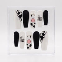 A set of 10 coffin-shaped press-on nails displayed in a clear acrylic case. The nails feature a black and white cow print, solid black, solid white with a pink cow face, and the word 'Moo' in black script.