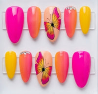 A set of almond glossy press-on nails displayed on a white background. The set features multiple almond-shaped nails in various colors: bright hot pink, peach-to-pink gradients, solid yellow, rhinestones and one nail with a pink and yellow butterfly illustration.
