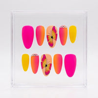 A set of 10 matte almond press-on nails displayed inside a clear acrylic case. The nails feature a variety of colors and designs, including solid hot pink, orange-to-yellow gradient nails, solid yellow nails, and two peach-toned nails with a pink and yellow butterfly print and rhinestone accents.