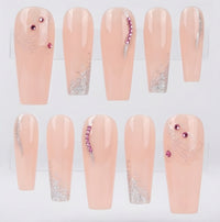 Ten coffin-shaped press-on nails displayed on a clear stand against a white background. The nails have a glossy pale peach-pink base with silver glitter swirl designs and decorative accents of pink rhinestones on two nails.