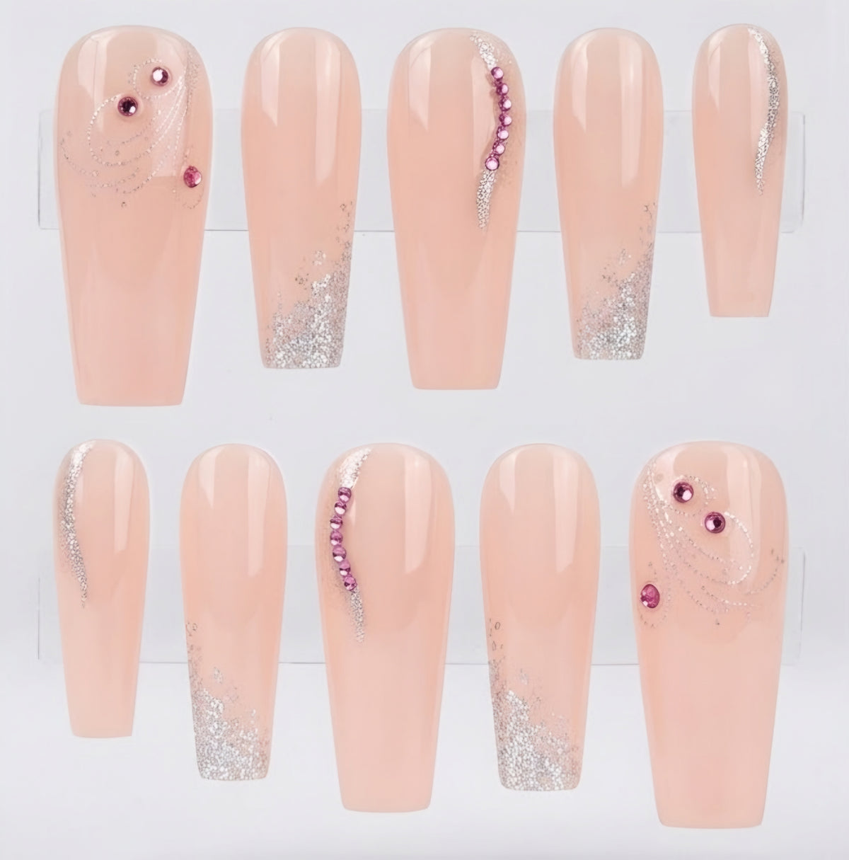 Ten coffin-shaped press-on nails displayed on a clear stand against a white background. The nails have a glossy pale peach-pink base with silver glitter swirl designs and decorative accents of pink rhinestones on two nails.