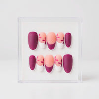 A set of 10 matte oval press-on nails displayed in a clear acrylic case against a white background. The nails feature a color-blocked design with deep burgundy, peach, and white, with some nails decorated with small red floral details.