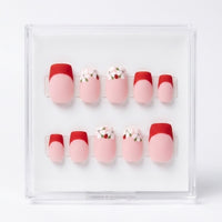 A set of 10 press-on nails displayed in a clear acrylic case against a white background. The nails feature a French tip design with a red half and a light pink full nail, with two additional designs featuring white flowers with ladybirds and green leaves.