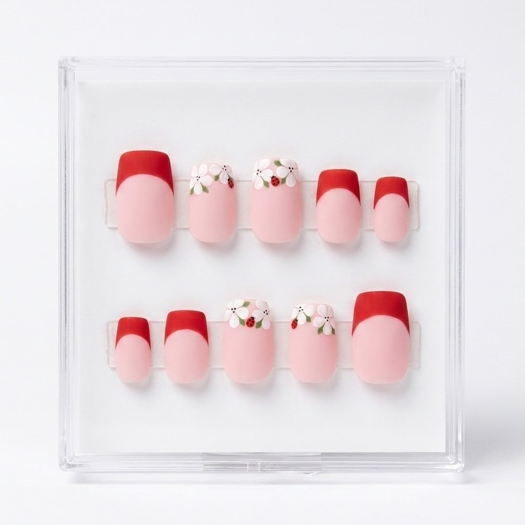 A set of 10 press-on nails displayed in a clear acrylic case against a white background. The nails feature a French tip design with a red half and a light pink full nail, with two additional designs featuring white flowers with ladybirds and green leaves.
