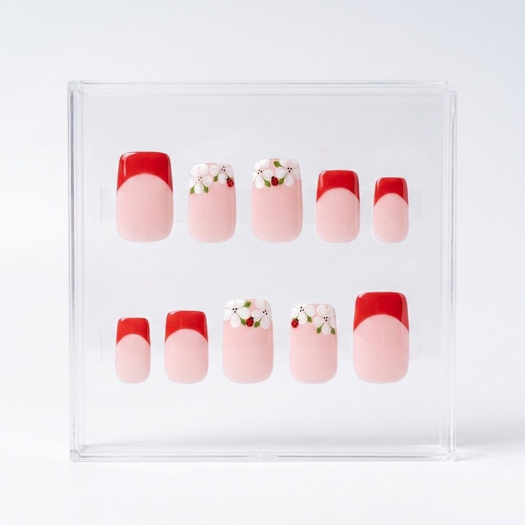 A set of 10 press-on nails displayed in a clear acrylic case against a white background. The nails feature a French tip design with a red half and a light pink full nail, with two additional designs featuring white flowers with ladybirds and green leaves.