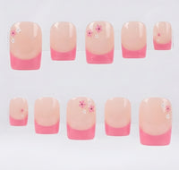 Ten square press-on nails arranged in two rows on a white background. The nails have a glossy nude-pink base and pink tips, with a few small floral accents in pink and white.