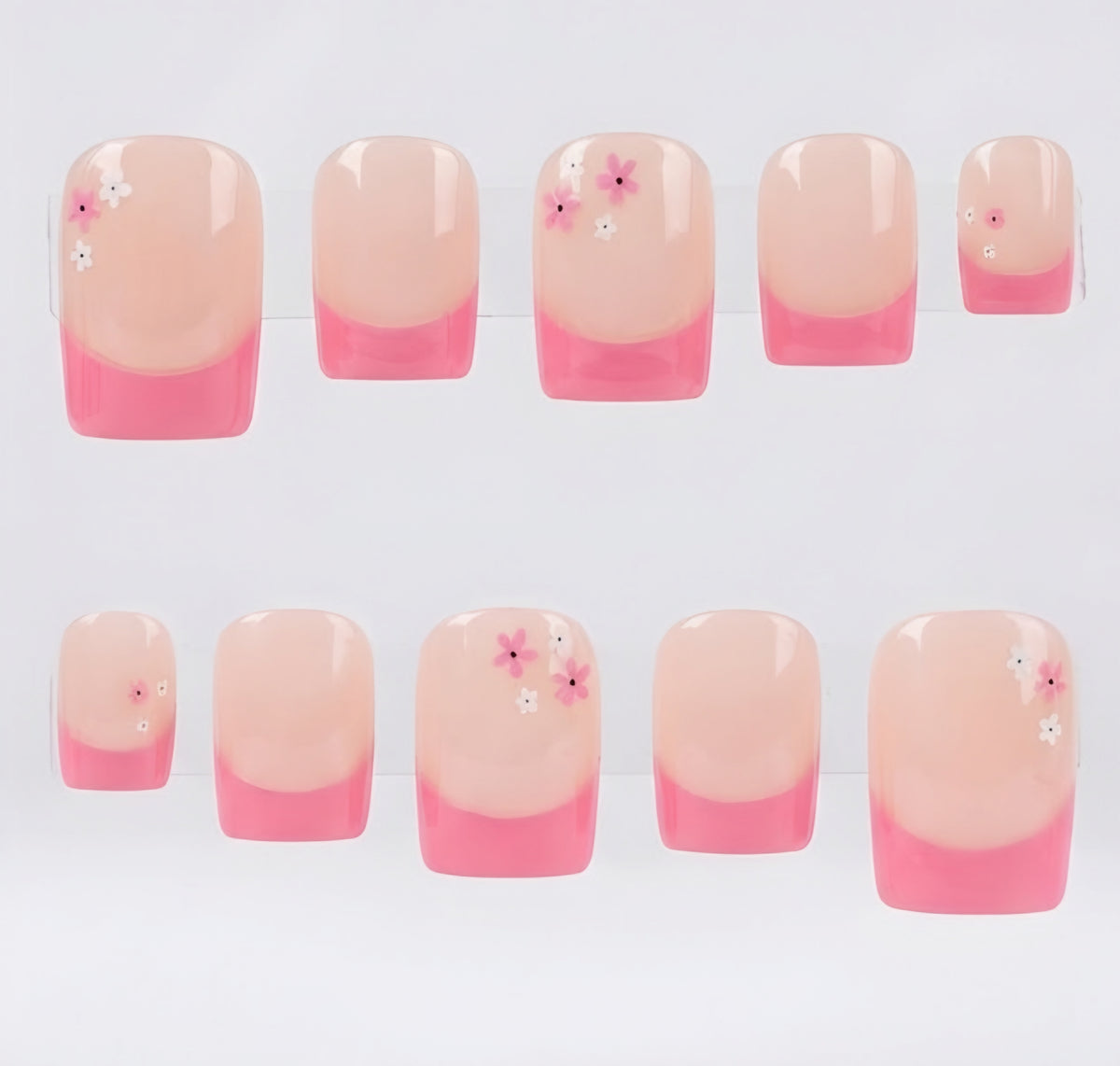 Ten square press-on nails arranged in two rows on a white background. The nails have a glossy nude-pink base and pink tips, with a few small floral accents in pink and white.