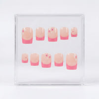 A set of ten square press-on nails displayed inside a clear acrylic case against a white background. The nails feature a glossy nude-pink base and a  pink french tip, with a few decorated with tiny pink and white flower details.