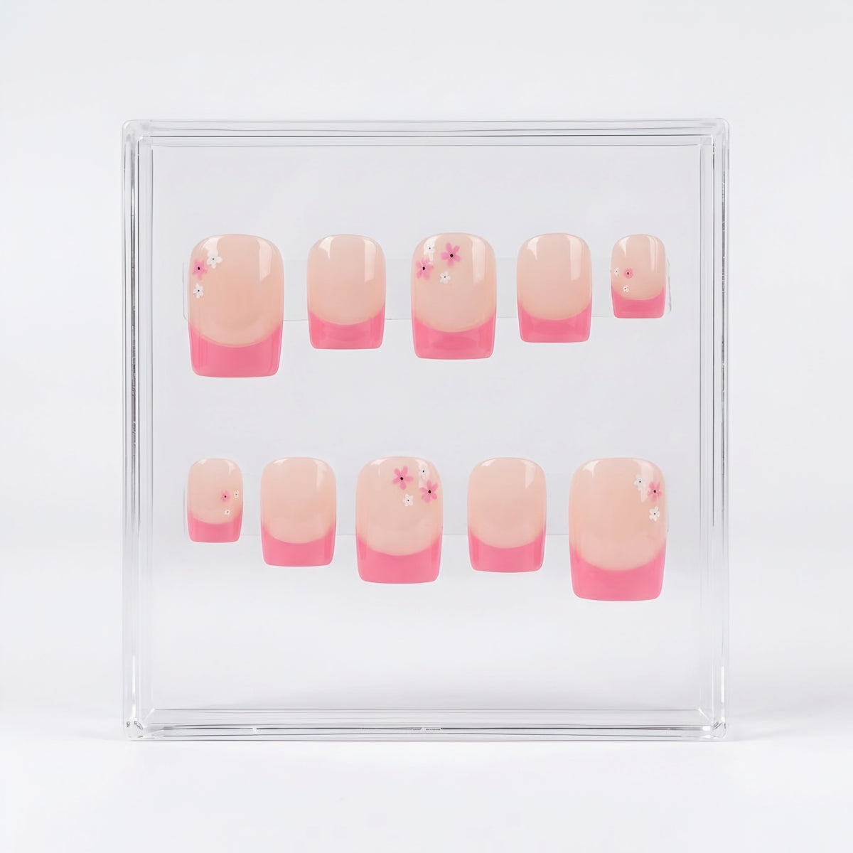 A set of ten square press-on nails displayed inside a clear acrylic case against a white background. The nails feature a glossy nude-pink base and a  pink french tip, with a few decorated with tiny pink and white flower details.