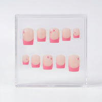 A set of ten square matte press-on nails displayed inside a clear plastic case on a white background. The nails feature a classic French manicure with a nude/pink base and  pink tips, with three nails decorated with tiny pink and white flower details.
