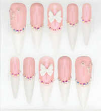 Ten stiletto press-on nails arranged in two rows on a white display card. The nails have a glossy pale pink base, a white  tips, and decorative accents including a white bow on two nails, small rhinestone clusters, and floral embellishments.