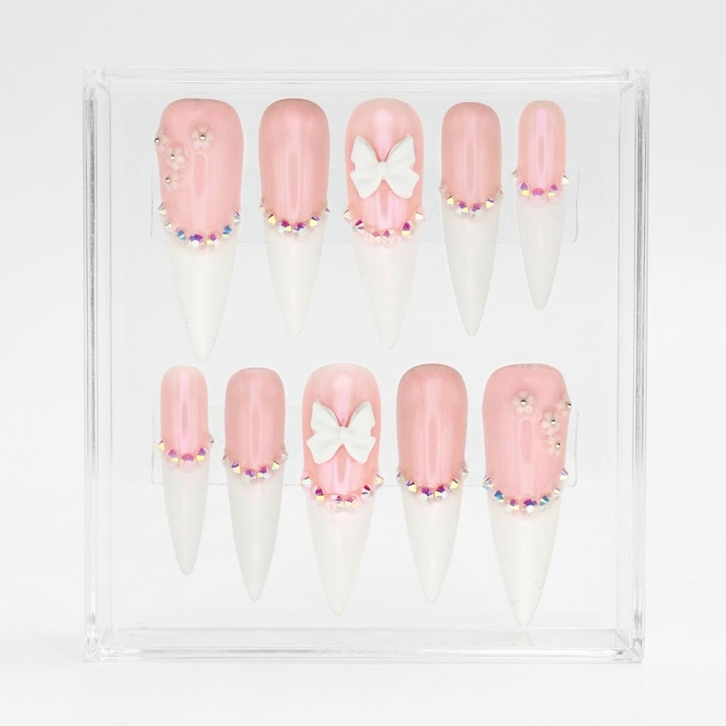 A set of 10 stiletto press-on nails displayed in a clear acrylic case against a white background. The nails feature a glossy light pink base, a white tip, and decorative details including a white bow, rhinestones, and small floral accents.