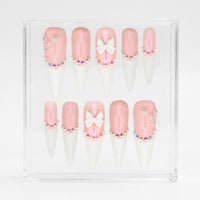 A set of 10 stiletto press-on nails displayed in a clear acrylic case against a white background. The nails feature a glossy light pink base, a white tip, and decorative details including a white bow, rhinestones, and small floral accents.