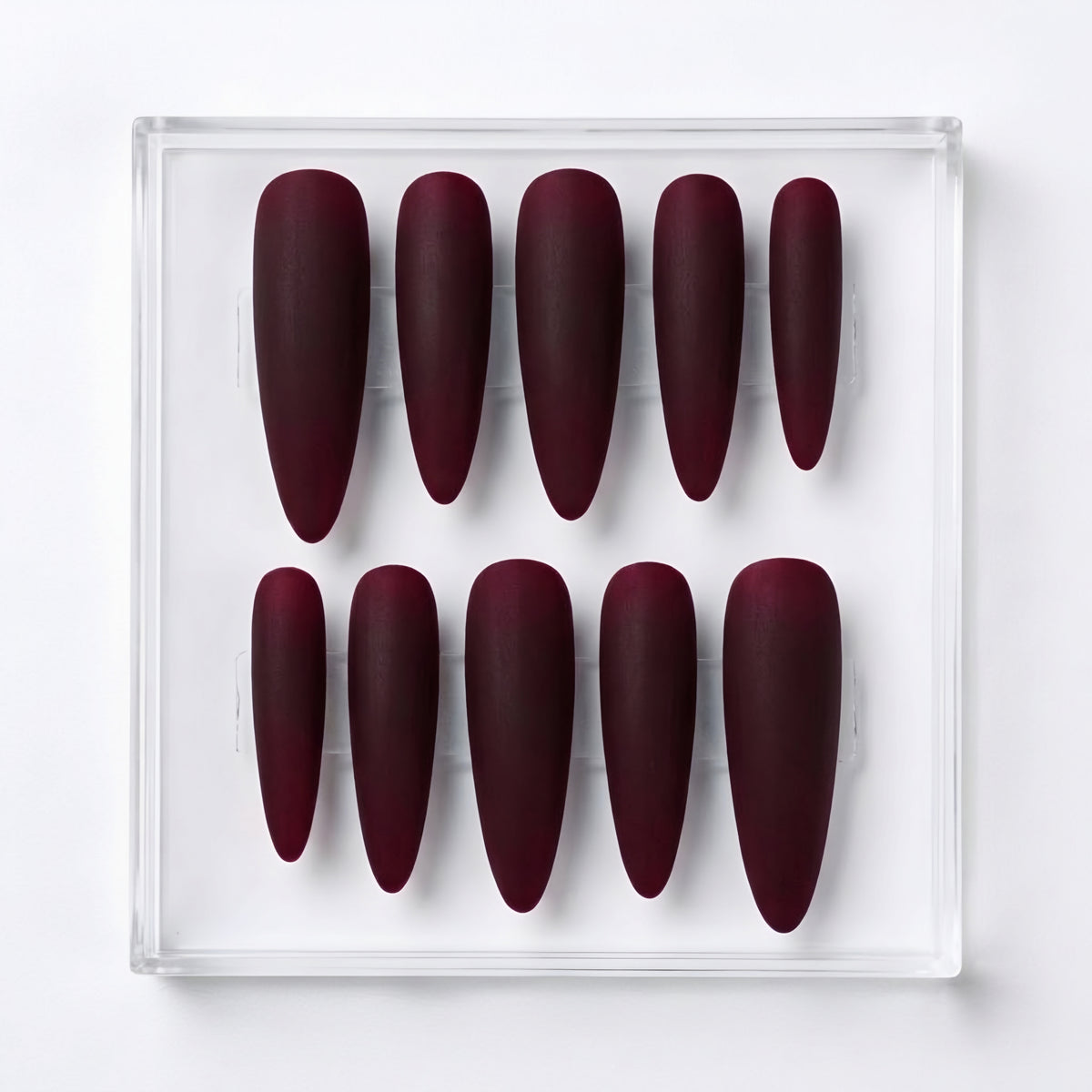 A set of ten long almond-shaped press-on nails in a deep matte burgundy color, arranged in two rows inside a clear acrylic display box on a white background.