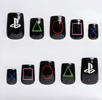 Ten black square glitter press-on nails arranged in two rows on a clear plastic display card against a white background. The nails feature PlayStation logo and controller button symbols in white, green, red, pink, and blue.