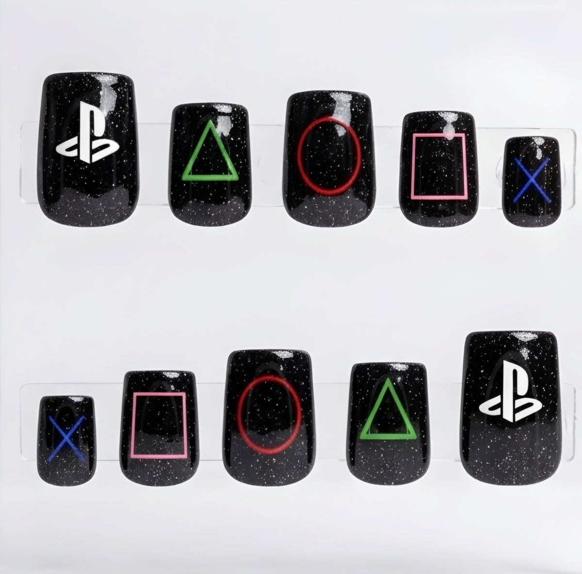 Ten black square glitter press-on nails arranged in two rows on a clear plastic display card against a white background. The nails feature PlayStation logo and controller button symbols in white, green, red, pink, and blue.