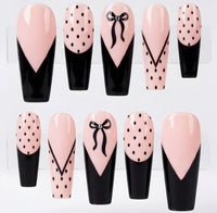 Ten coffin-shaped press-on nails arranged in two rows on a white background. The nails feature a glossy pale pink and black design with some decorated in black polka dots, and one with a black bow motif.