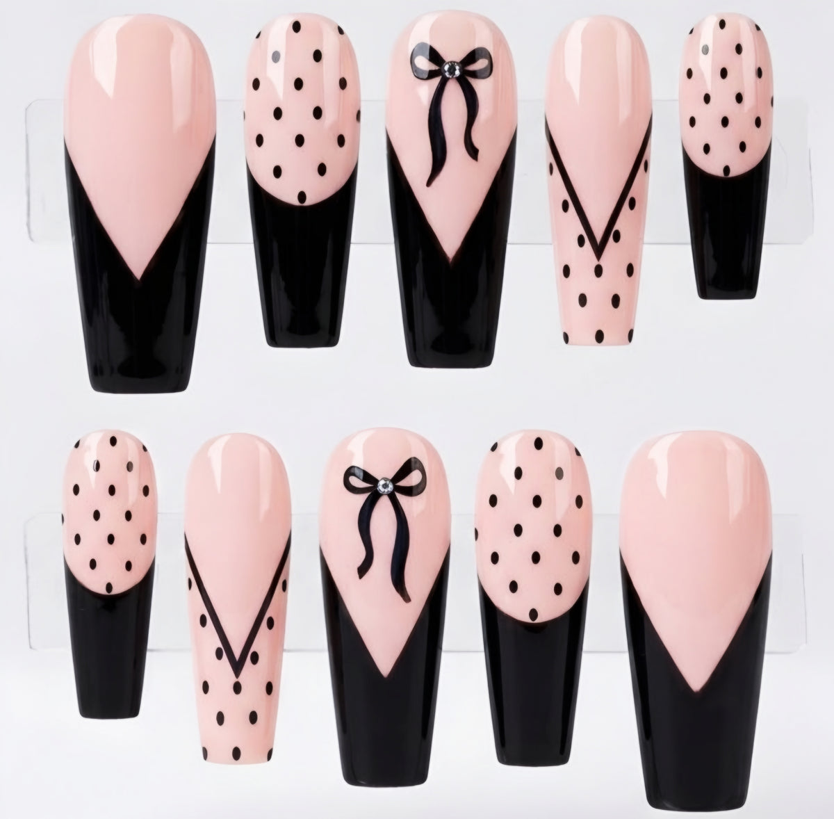 Ten coffin-shaped press-on nails arranged in two rows on a white background. The nails feature a glossy pale pink and black design with some decorated in black polka dots, and one with a black bow motif.