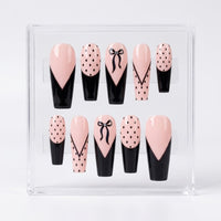A set of 10 coffin  shaped glossy press-on nails displayed in a clear acrylic case against a white background. The nails feature a design of light pink and black with various patterns including polka dots, bow details, and lines.