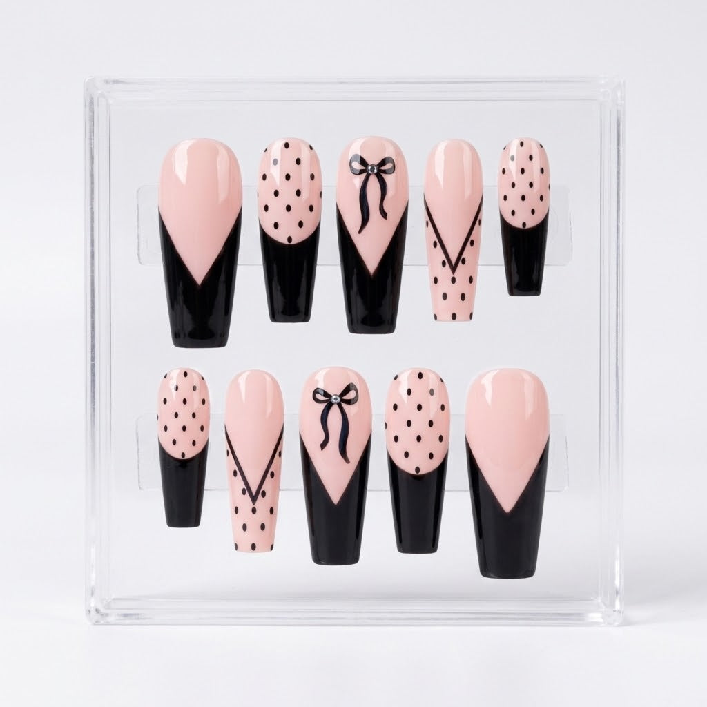 A set of 10 coffin  shaped glossy press-on nails displayed in a clear acrylic case against a white background. The nails feature a design of light pink and black with various patterns including polka dots, bow details, and lines.