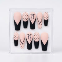 A set of 10 matte coffin shaped press-on nails displayed in a clear acrylic case against a white background. The nails feature a black and pale pink color scheme with various patterns including polka dots, lines, french tips and bows.