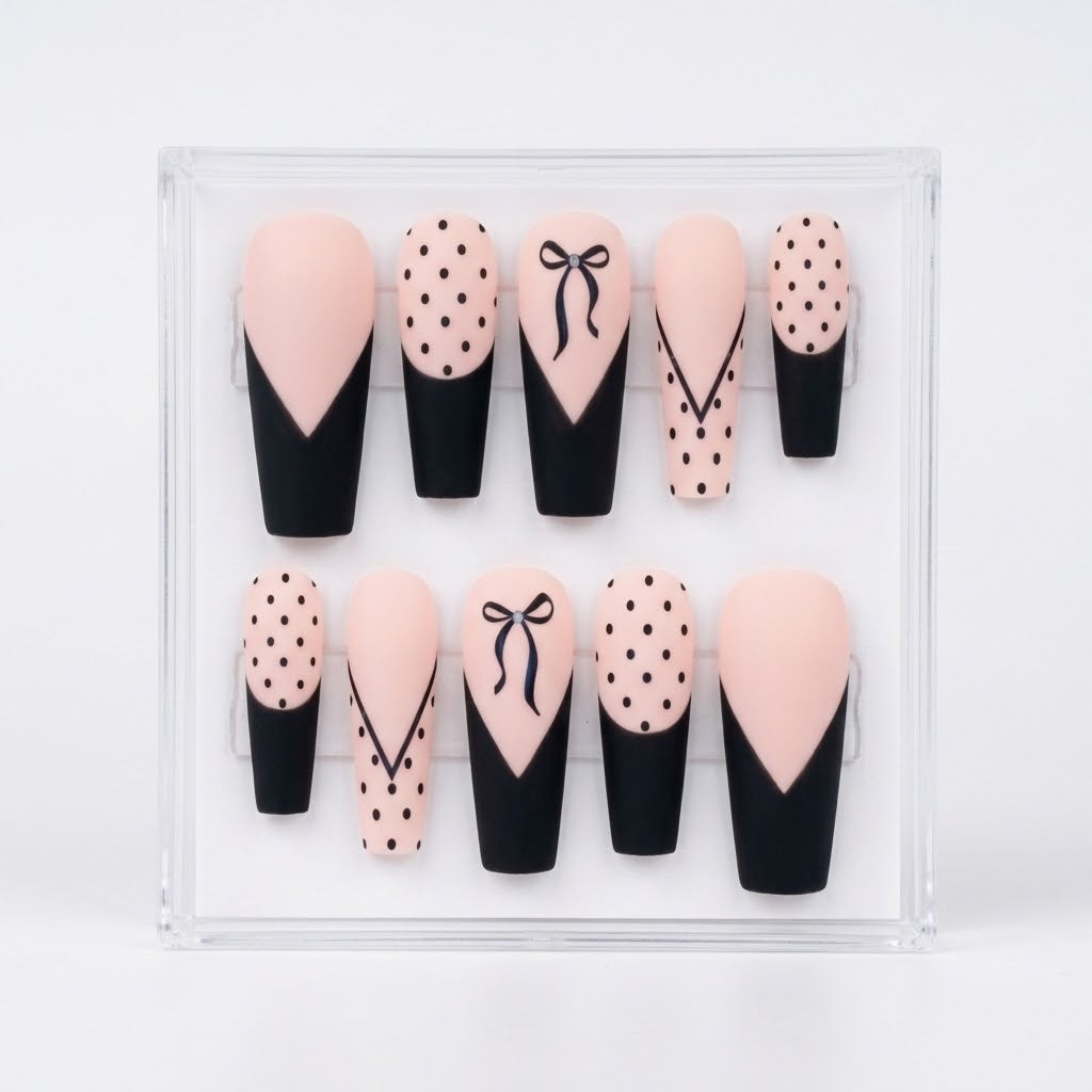 A set of 10 matte coffin shaped press-on nails displayed in a clear acrylic case against a white background. The nails feature a black and pale pink color scheme with various patterns including polka dots, lines, french tips and bows.