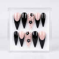 A set of 10 false press-on nails displayed in a clear plastic case against a white background. The nails feature a mix of glossy black stiletto shapes, glittery pale pink stiletto shapes, and pale pink almond-shaped nails decorated with black floral motifs and polka dots.