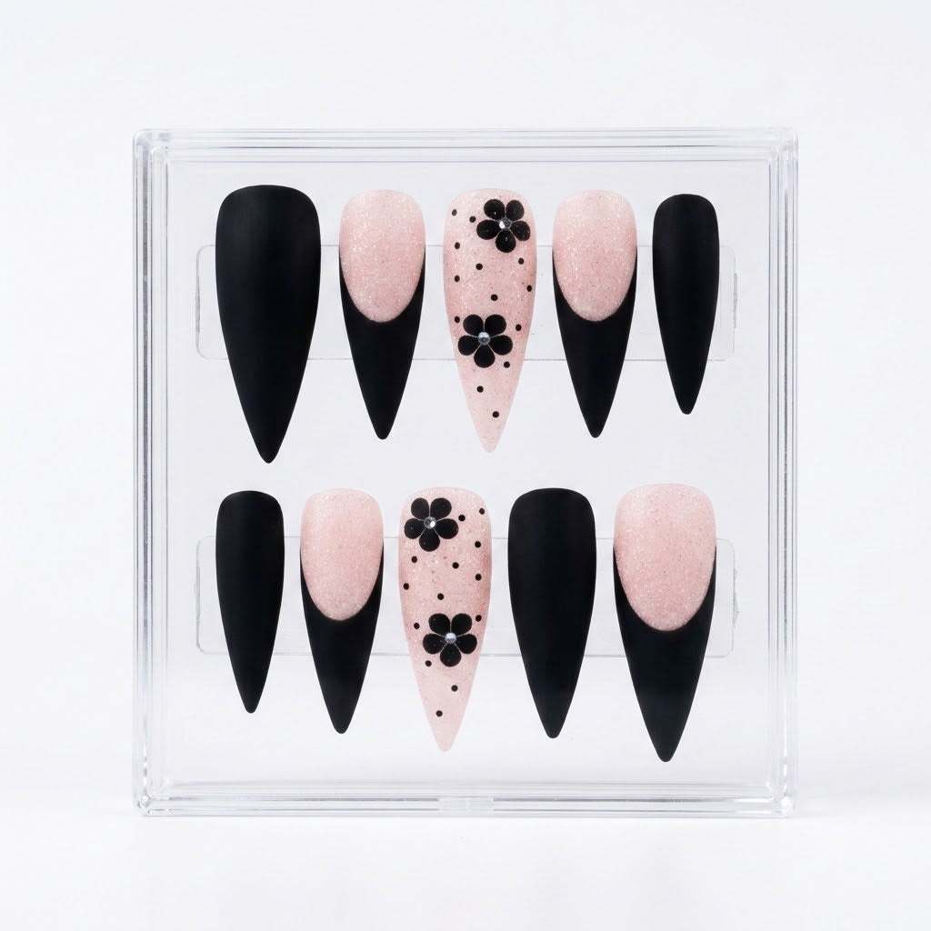 A set of 10 press-on nails in a clear plastic display box, featuring a black and pale pink color scheme with two nails decorated with black flowers and polka dots.
