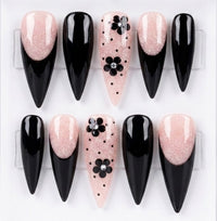 Ten stiletto press-on nails arranged in two rows on a white background. The design alternates between glossy black nails and pale pink glitter nails, with one center nail featuring a black flower motif with small silver rhinestones.