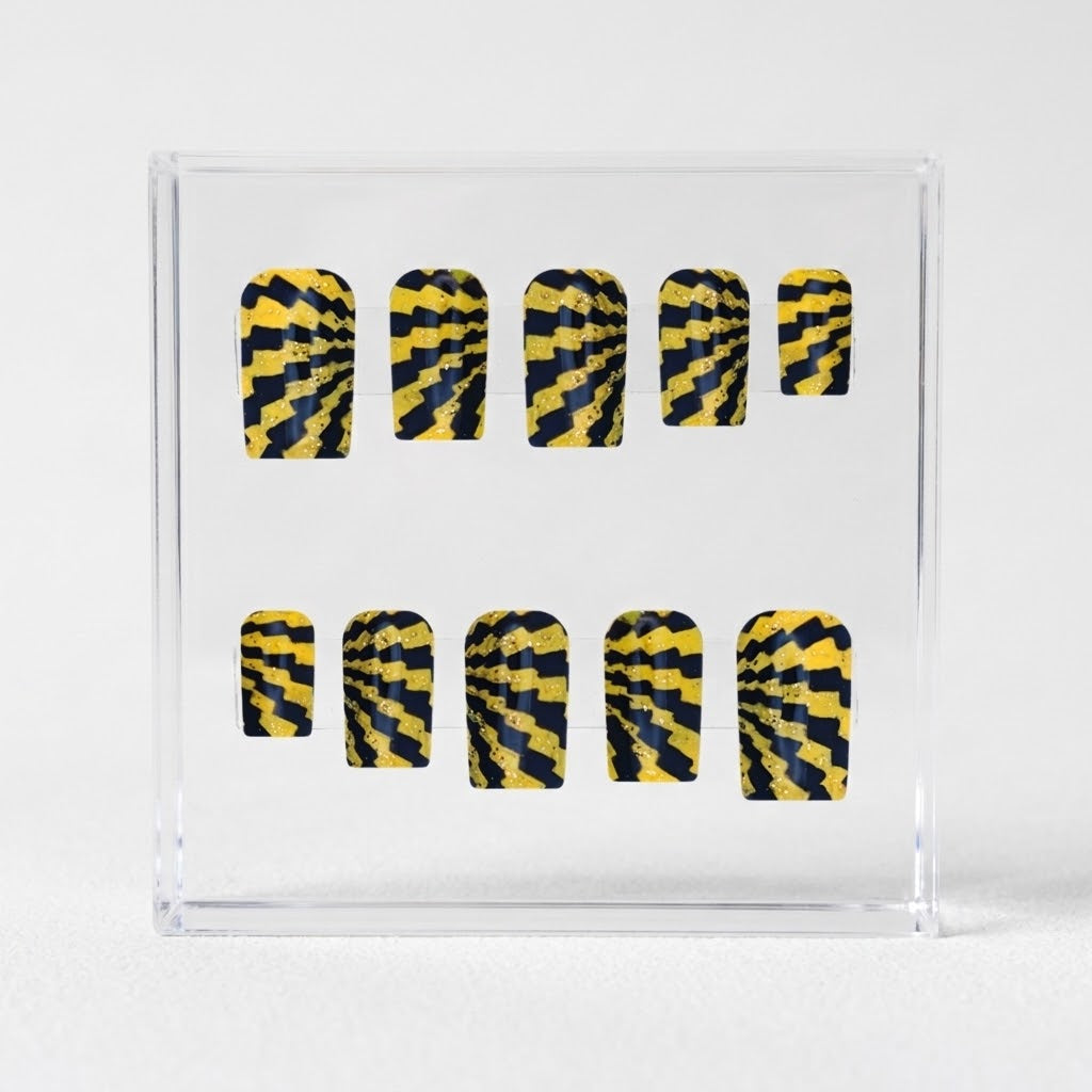 A set of ten press-on glossy nails with a yellow and black spiral stripe pattern, displayed inside a clear acrylic block on a light gray background.