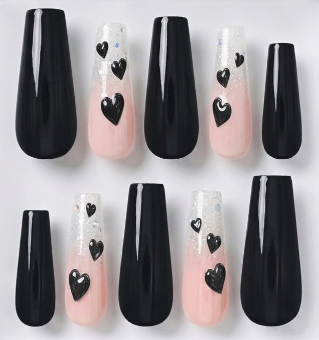 A set of long, coffin-shaped press-on nails arranged in two rows on a plain white background. The nails feature three colorways: glossy black, a soft pink-to-white glitter ombre gradient with black heart decals, and a white-to-pink ombre gradient with black heart decals and subtle glitter.