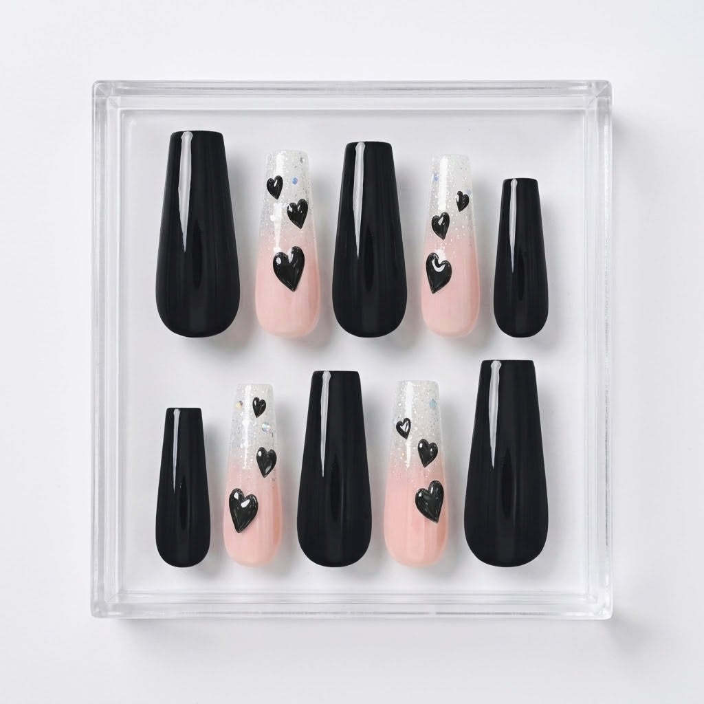 A set of coffin-shaped press-on nails displayed in a clear acrylic case on a light gray background. The nails feature a glittery white and pale pink ombre design with small black heart details on some of the tips.