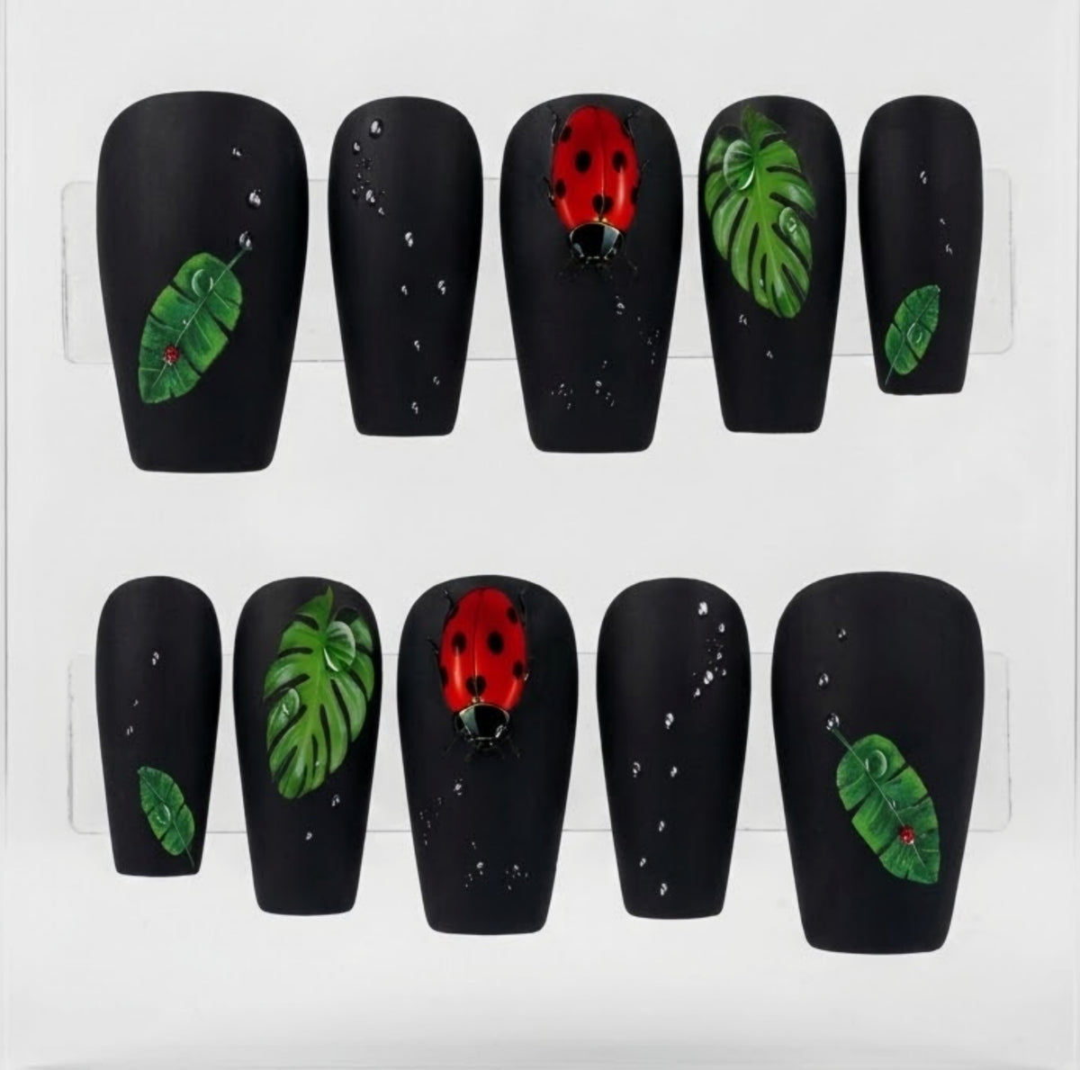 A set of coffin-shaped press-on nails displayed on a white background. The nails are matte black with various decorative designs, including green monstera leaves, small green leaves, water drops, and a red ladybug with black spots.
