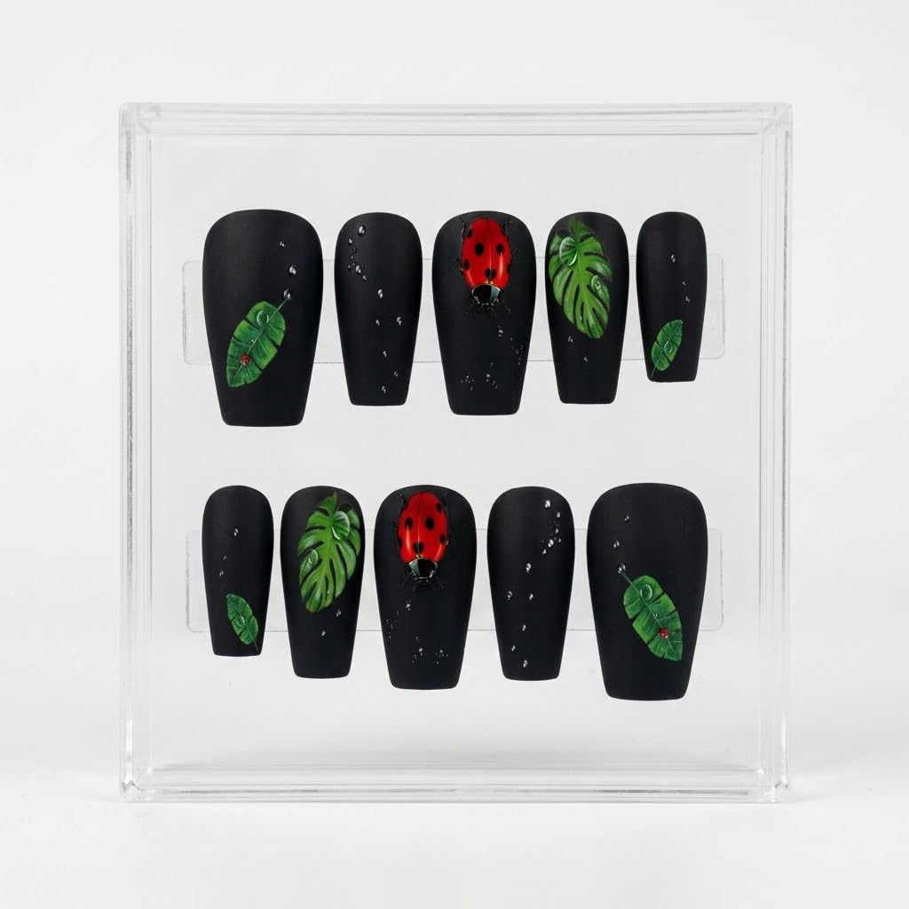 A set of 10 coffin shaped black press-on nails displayed in a clear acrylic box. The nails feature green tropical leaf designs and red ladybug motifs with small clear water drops.