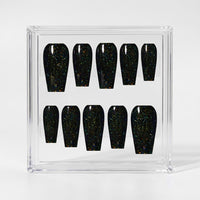 Ten coffin-shaped press-on nails with a glossy black finish and fine iridescent multicoloured glitter, displayed inside a clear acrylic case on a light gray background.