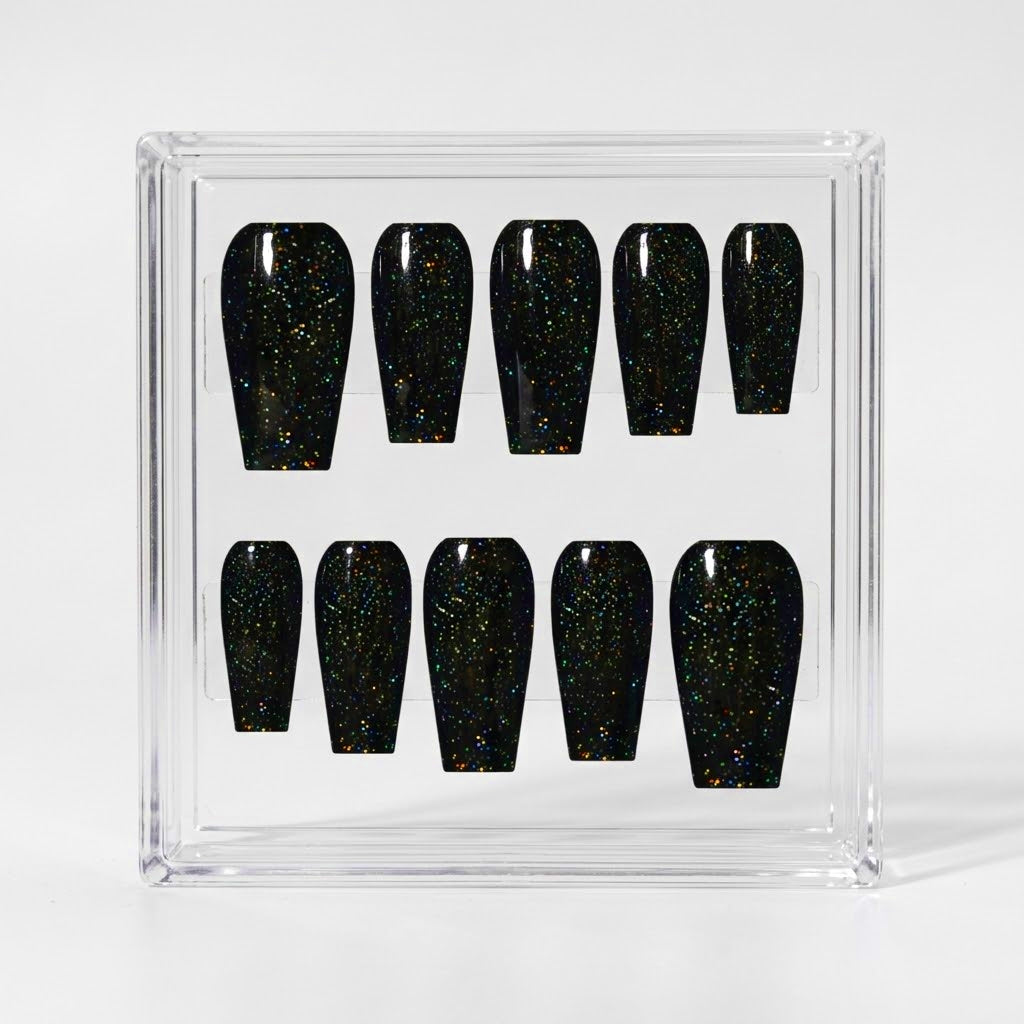 Ten coffin-shaped press-on nails with a glossy black finish and fine iridescent multicoloured glitter, displayed inside a clear acrylic case on a light gray background.