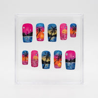 A set of 10 square press-on nails displayed inside a clear plastic square box on a light gray background. The nails feature vibrant tropical beach scenes with pink, orange, yellow, and blue sunset gradients, black palm tree silhouettes, and glittery blue water details.
