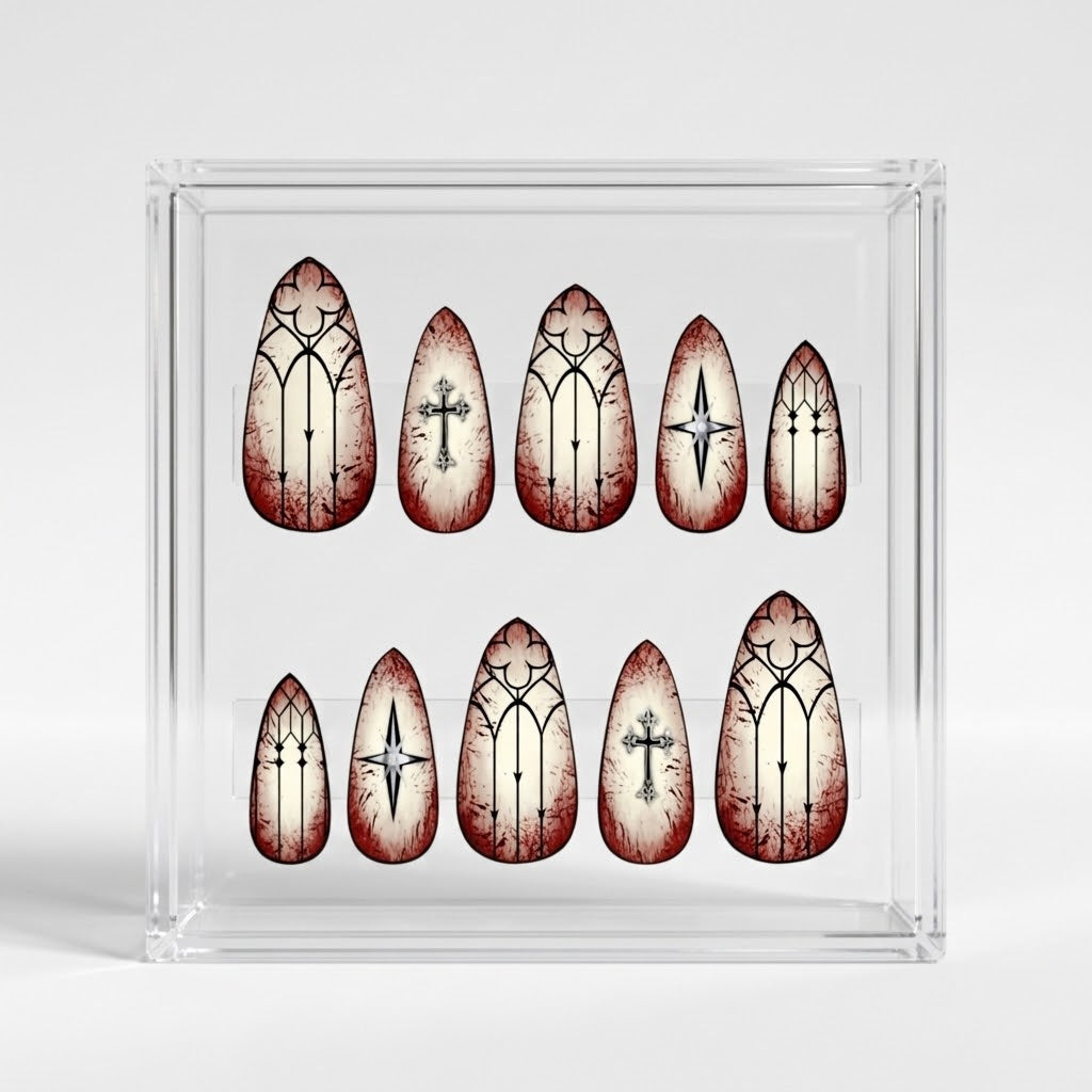 A set of almond shaped press-on nails displayed inside a clear acrylic case on a light gray background. The nails feature a distressed off-white background with dark red blood-like splatters and gothic black line art, including crosses and arched window designs.