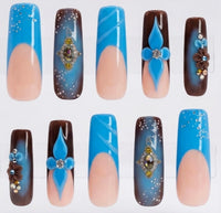 Ten false press-on nails arranged in two rows on a plain white background. The nails feature various designs including solid bright blue, a blue and nude-peach split design, dark brown with blue floral accents, and dark brown with blue gradient and gem details.