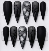 Ten Stiletto-shaped false nails displayed on a white card, featuring matte black and gray gothic designs including silhouettes of a figure, bare trees, and flying bats.