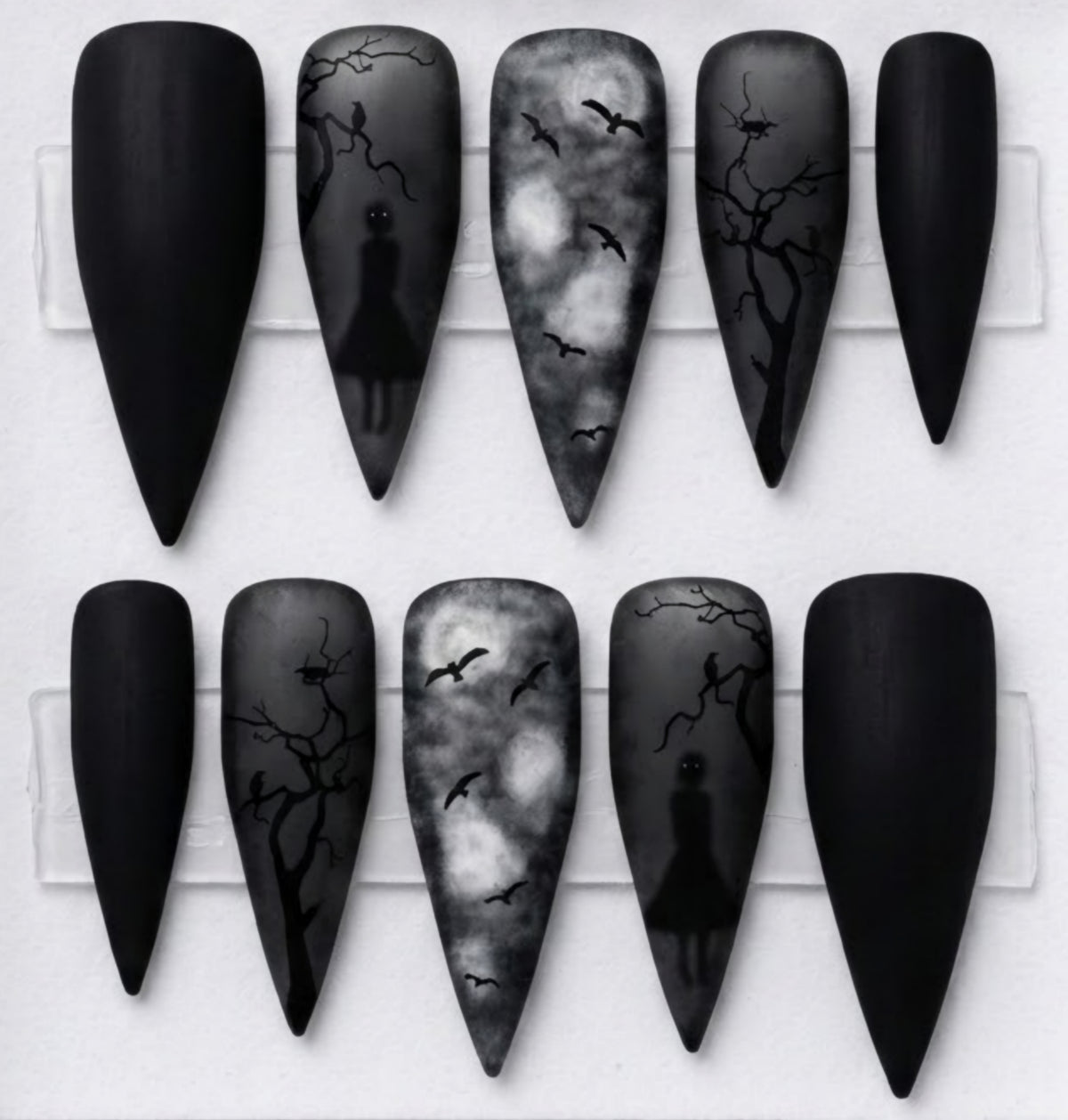 Ten Stiletto-shaped false nails displayed on a white card, featuring matte black and gray gothic designs including silhouettes of a figure, bare trees, and flying bats.