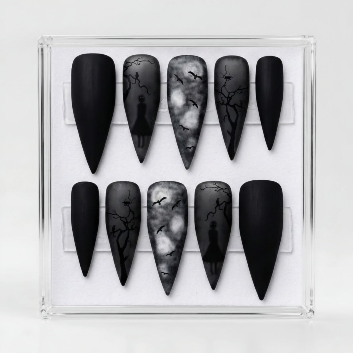 A set of stiletto-shaped press-on nails displayed in a clear acrylic case. The nails feature a gothic theme with matte black, smoky gray, and white designs including silhouettes of figures and bare tree branches.