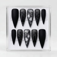 A set of stiletto-shaped press-on nails displayed in a clear acrylic case. The nails feature a gothic theme with matte black, smoky gray, and white designs including silhouettes of figures and bare tree branches.