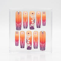 A set of ten square press-on nails displayed inside a clear square plastic case on a white background. The nails feature a warm orange-to-purple gradient design with white floral patterns, small pink flowers, and rhinestone embellishments.