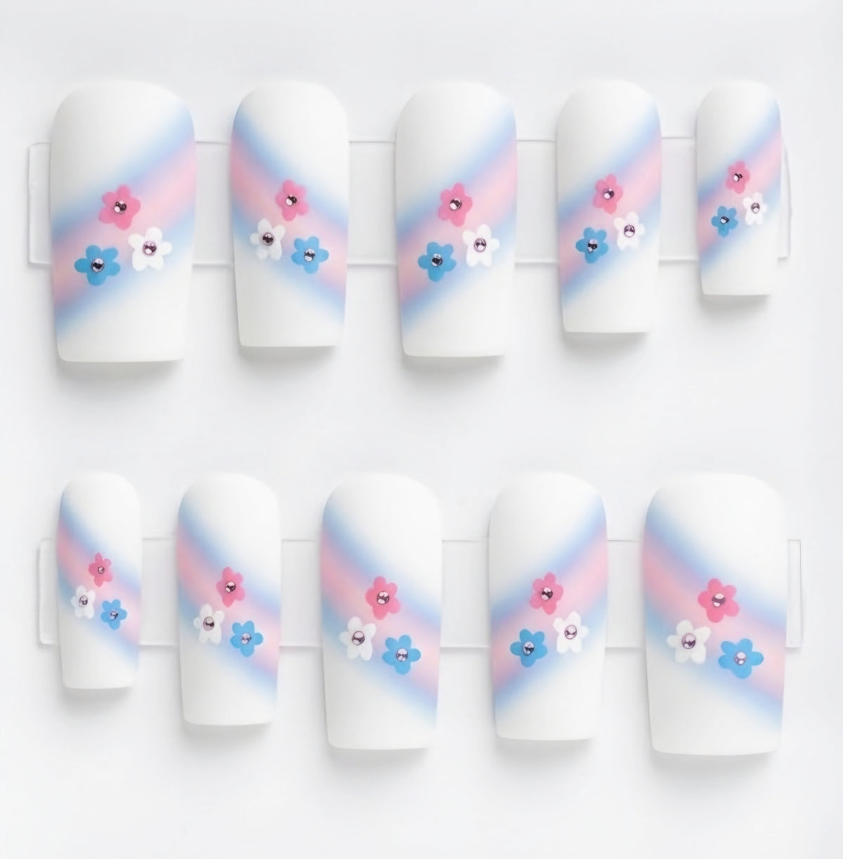 A set of 10 square white glossy press on nails featuring a trans colours themed design. With pink and pale bue ombre swipe and small pink, blue and white flowers with rhinestone centres.