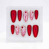 A set of 10 almond  press-on nails displayed in a clear acrylic case on a white background. The set features alternating red glitter and white decorated with red hearts and branch patterns.