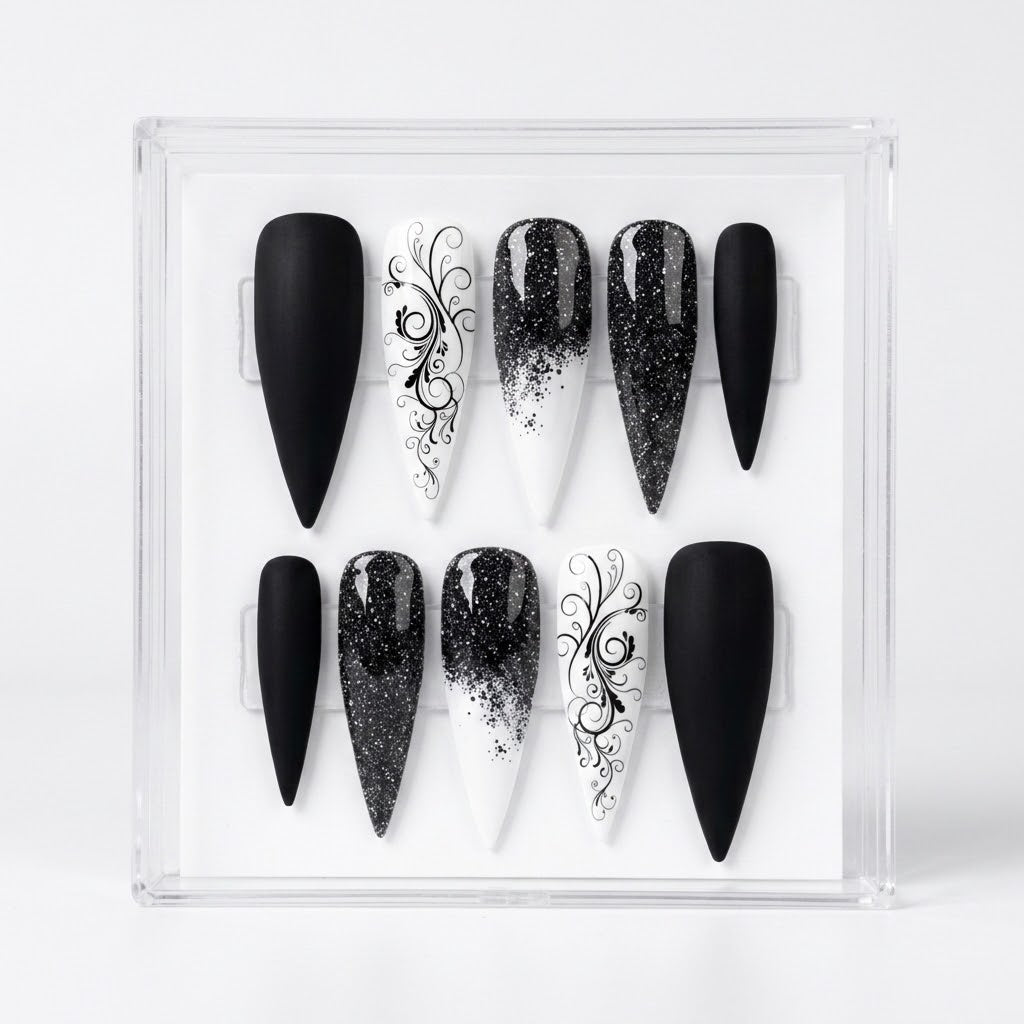 A set of 10 long Stiletto-shaped press-on nails displayed in a clear acrylic case against a white background. The nails feature black, white, and black-and-white gradient designs with glitter and elegant black scrollwork patterns.