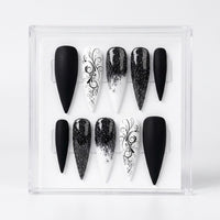 A set of 10 long Stiletto-shaped press-on nails displayed in a clear acrylic case against a white background. The nails feature black, white, and black-and-white gradient designs with glitter and elegant black scrollwork patterns.
