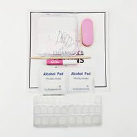Nail kit contents laid out on a white printed card over a speckled gray countertop, including a clear plastic tray, a pink nail file, a wooden stick, a pink tube labeled Nail Glide, two Alcohol Pad packets, and a sheet of adhesive nail tabs.